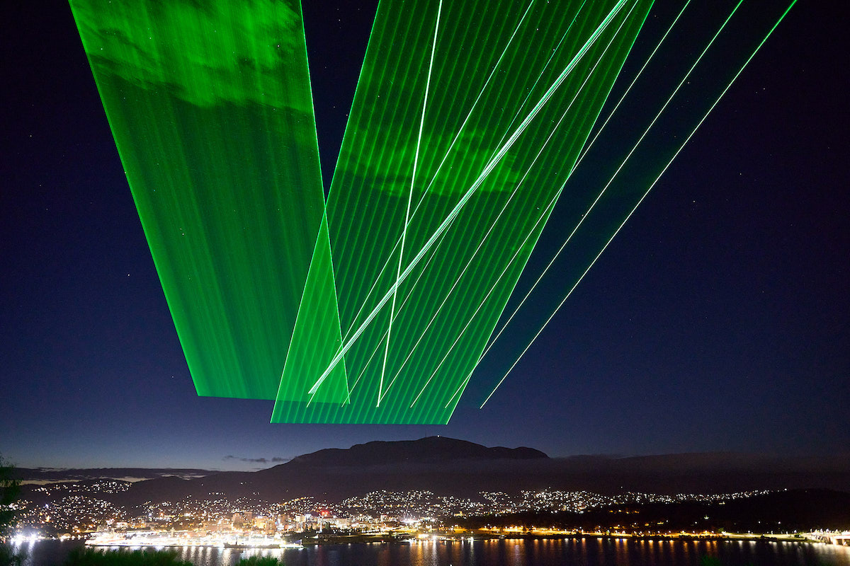 Planes of green laser lights over a mountain top, with beams cutting through clouds.