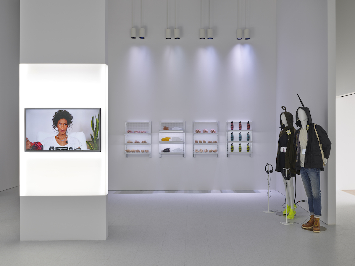 A view of a white gallery with a screen showing a digitized Whitney Houston, racks of faux products, and two mannequins wearing stylish outdoors fashions.